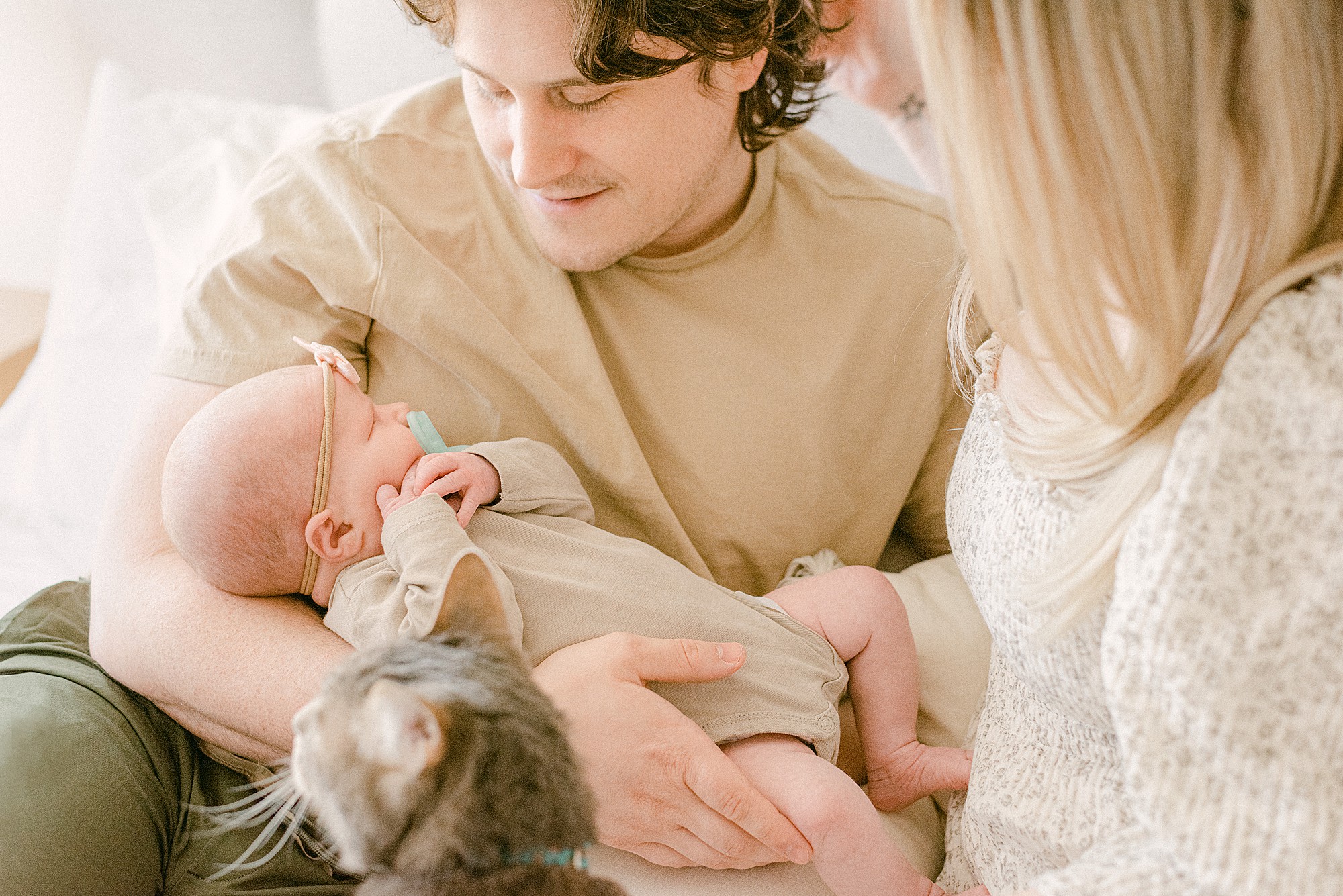 Gilbert Newborn photographer captures parents are holding newborn baby while sitting on bed. A grey cat pokes it's head in frame looking at baby.