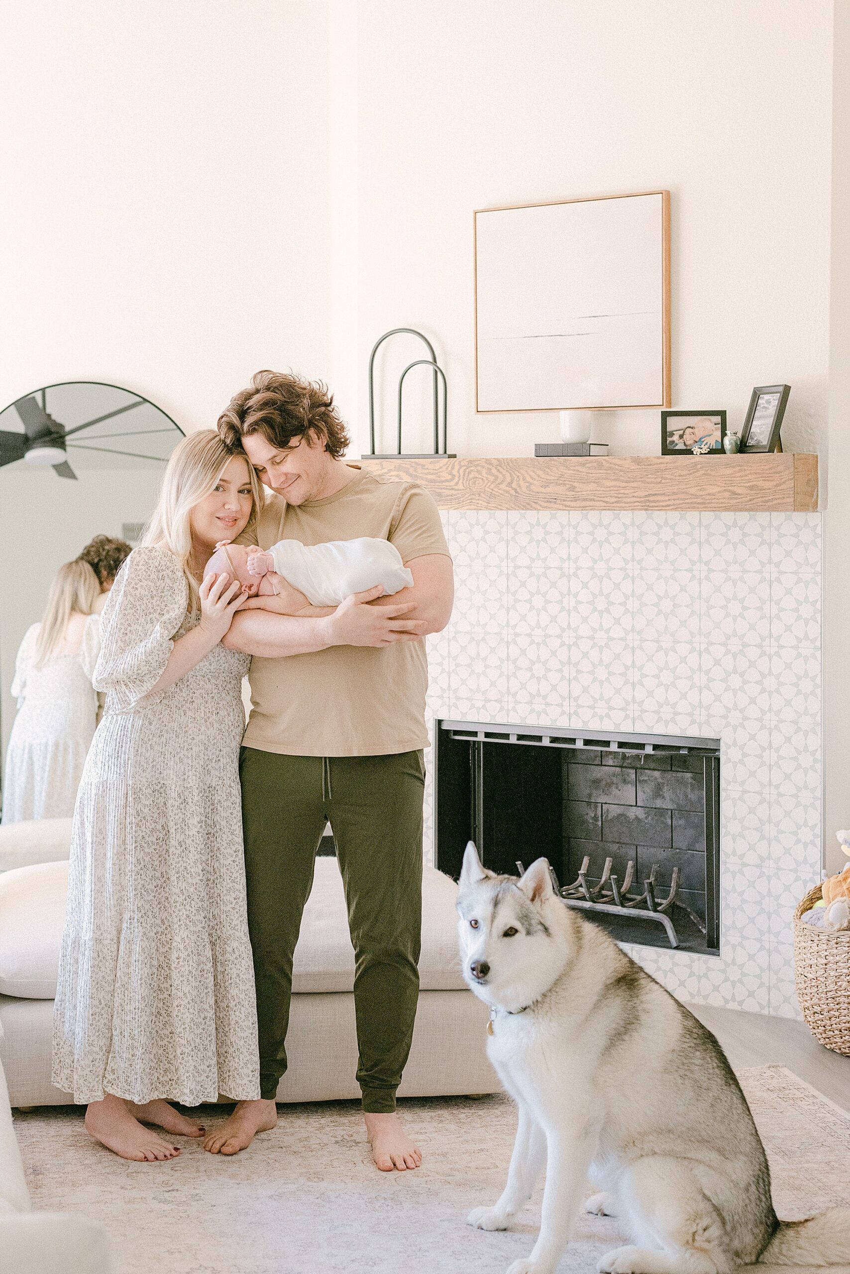 New parents are holding newborn baby in neutral decor Spanish styled home in Scottsdale AZ. Siberian Huskey is sitting on hind legs posed next to them.