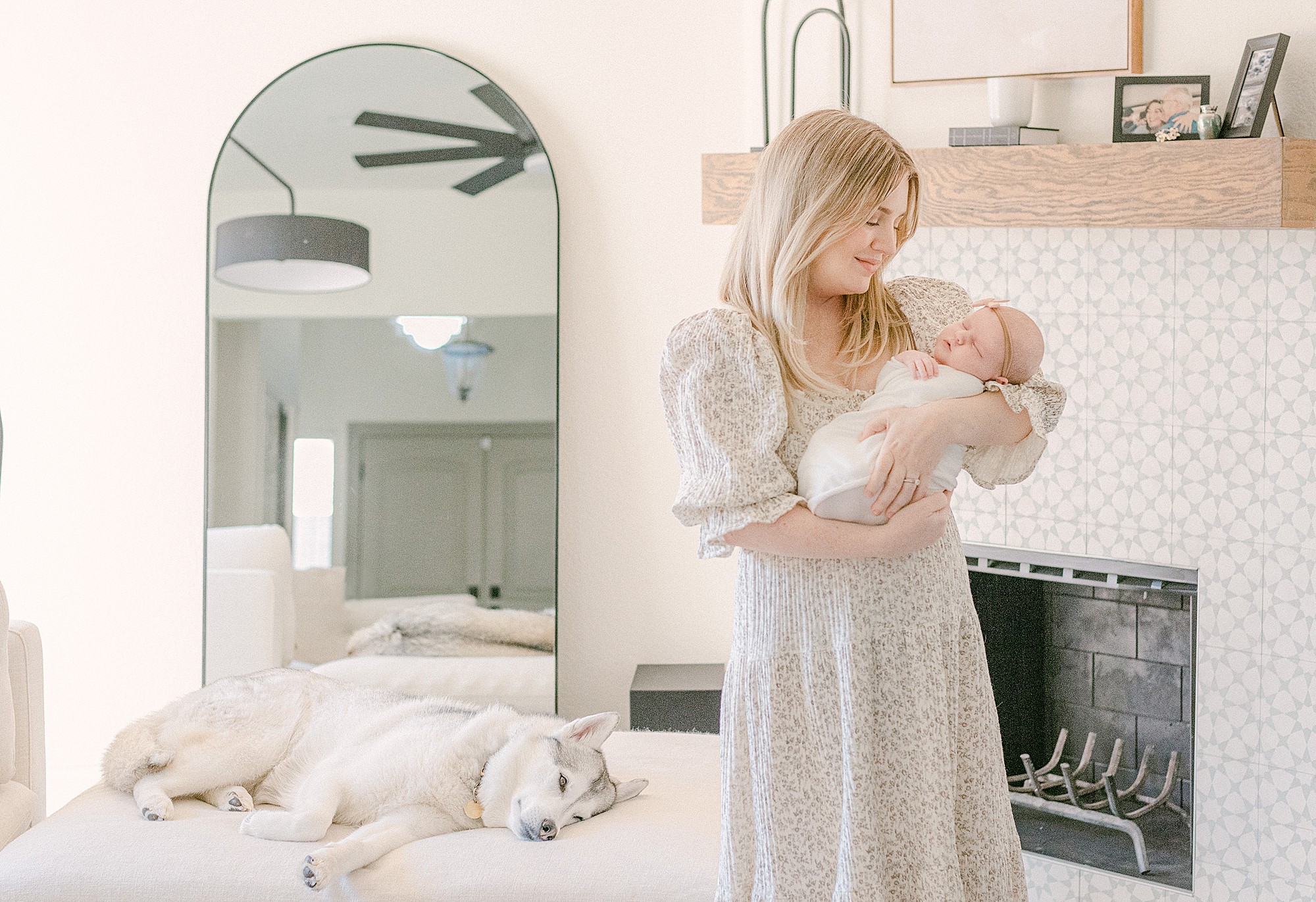 New mom holding baby in living room for in-home newborn photoshoot. Siberian Husky is laying on ottoman behind her.