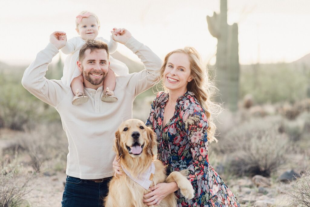 Family of three posing with dog together in the desert in the East Valley, AZ. They're surrounded by saguaro cactus and mountains in the background.