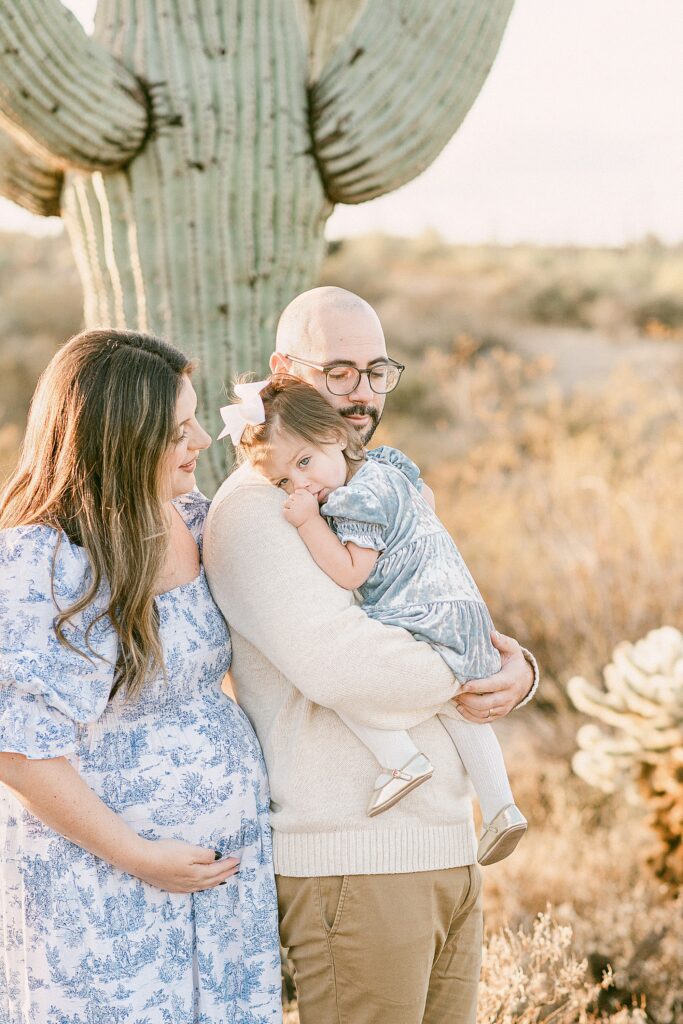 Family of three with toddler girl embraced for fall family photos in the desert with saguaro cactus. Mom is wearing blue and white dress, daughter in velvet blue dress with knit tights and ballet flats, and dad in cream sweater with brown chinos.