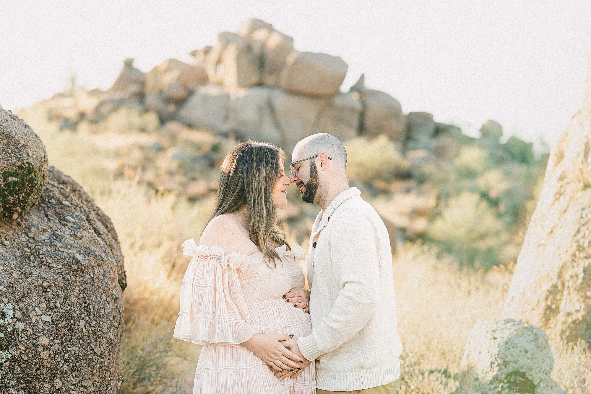 Expecting mom and dad face to face with hands on baby bump. Mom is wearing cream chiffon dress. Dad is in cream sweater and brown kahkis. They're styled in perfect outfits for fall family photos