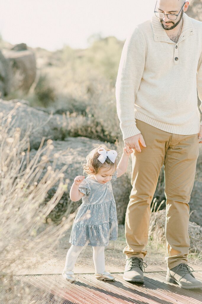 Dad wearing cream sweater and brown chinos holding hands and walking with toddler daughter. Daughter is wearing blue velvet dress, knit white tights, and gold ballet flats. They're styled as inspiration as perfect outfits for your fall family photos