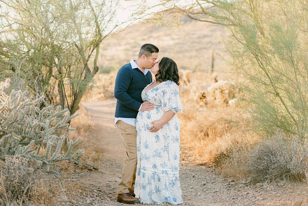 Couple standing under desert trees kissing while holding baby bump.