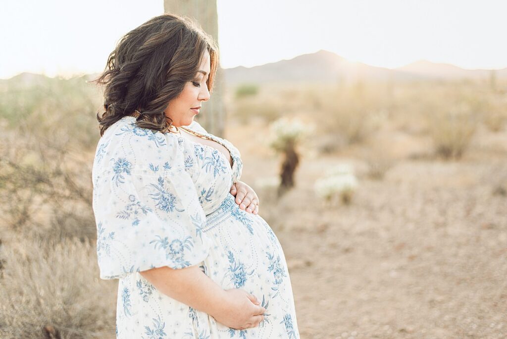 Expecting mom holding and looking down at baby bump. Desert mountain view is behind her with golden hour light.