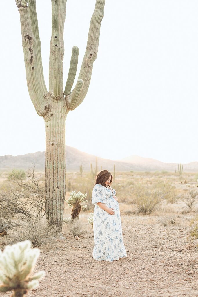 Pregnant woman standing in Phoenix desertscape. She's dressed in a flowy white dress next to saguaro cactus