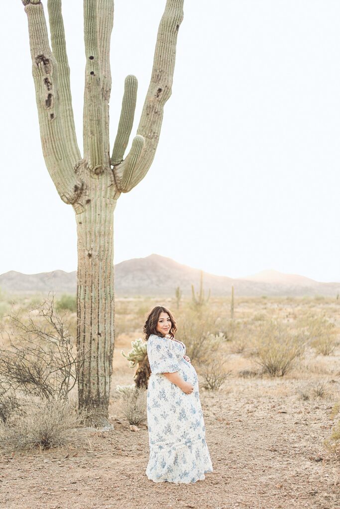 Pregnant woman holding baby bump and looking at camera. She's next to a tall saguaro cactus with golden hue mountains behind her