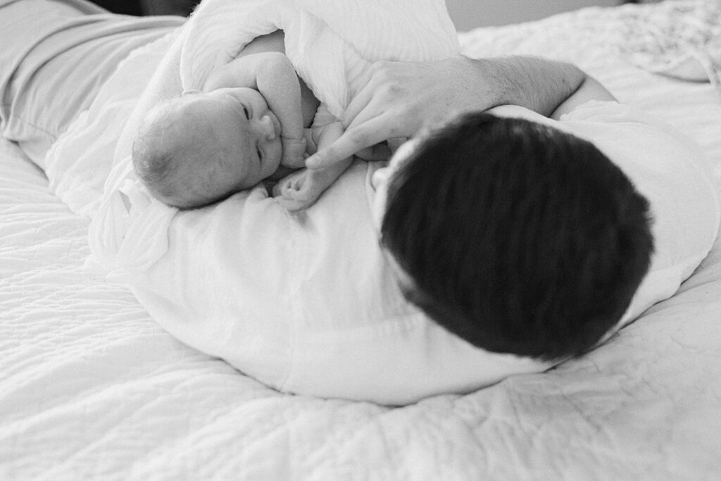 Dad is in relaxed position laying on his back on bed while holding newborn baby facing him.