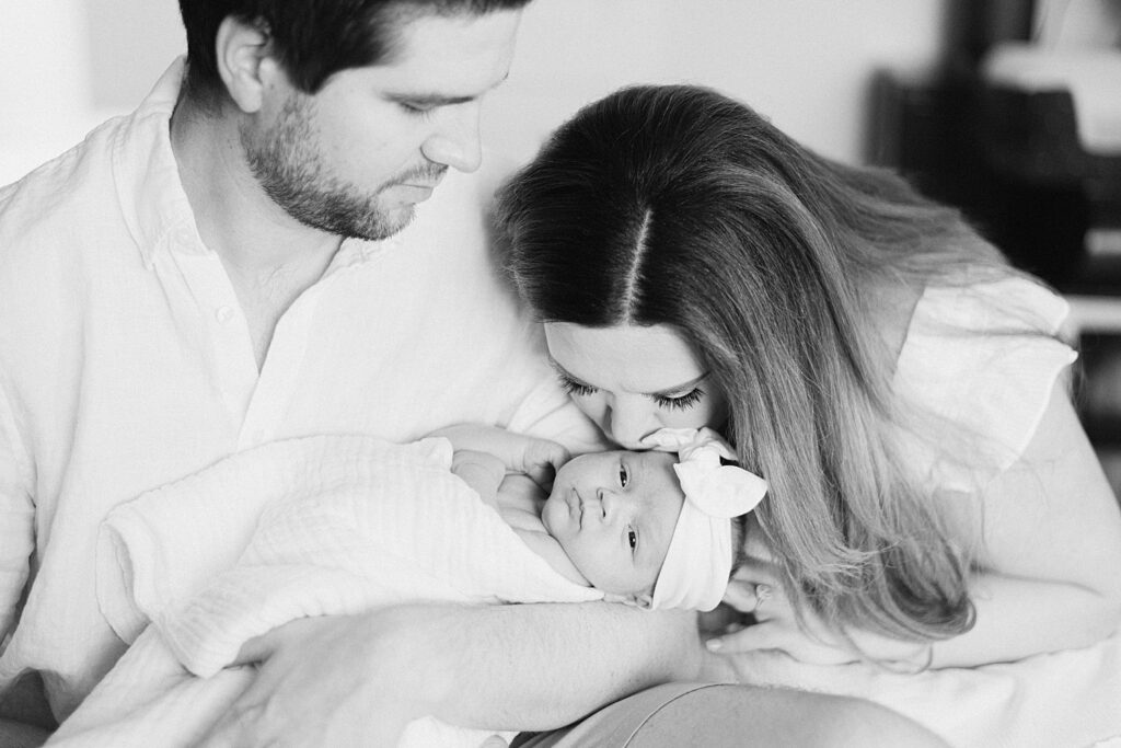 newborn baby is relaxed and content in dad's arms while mom is kissing baby's forehead