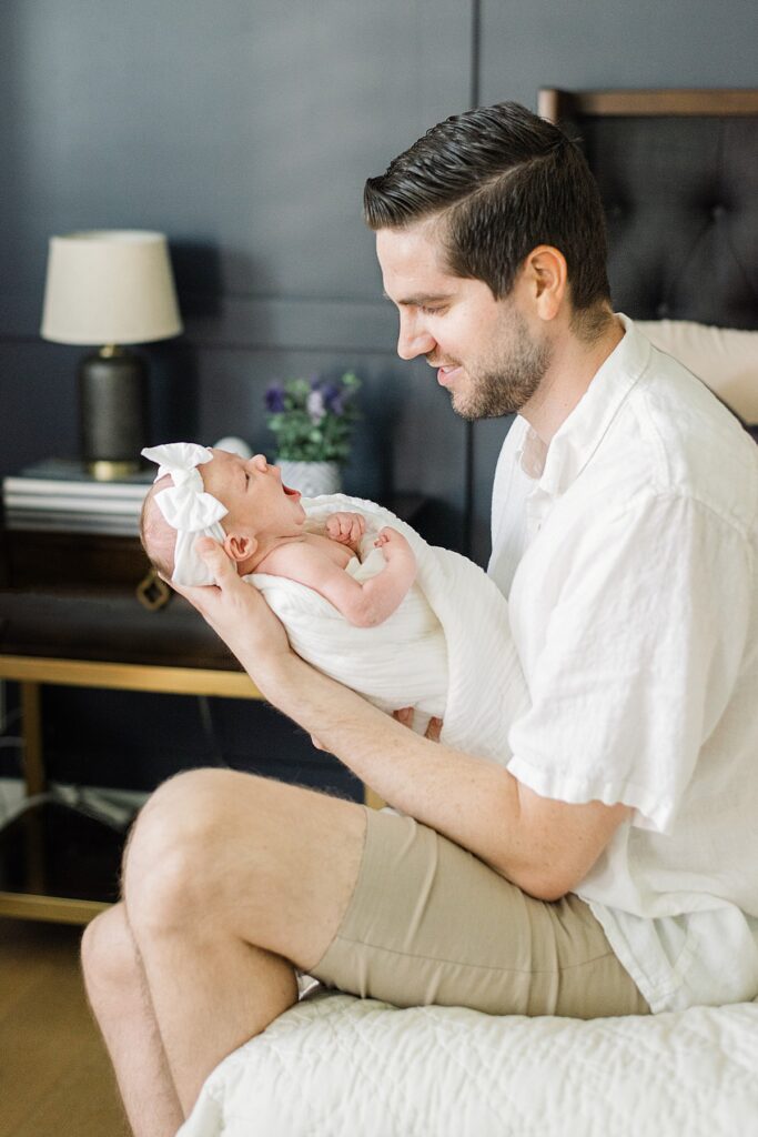 dad sitting on edge of bed while holding and looking at newborn