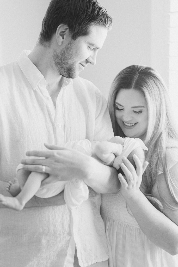 dad is holding newborn baby and smiling at mom while mom is looking at and smiling down at baby