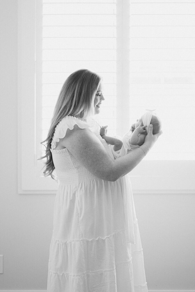 first time mom standing next to window wearing white hill house dress. she's cradling newborn baby in front of her