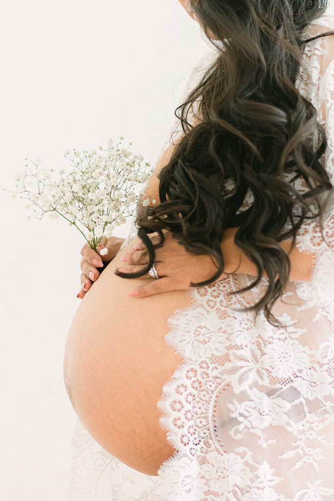 mom holding baby's breath flowers near baby bump. She's wearing a white queen anne's lace robe and her waist-length hair is covering her upper body.