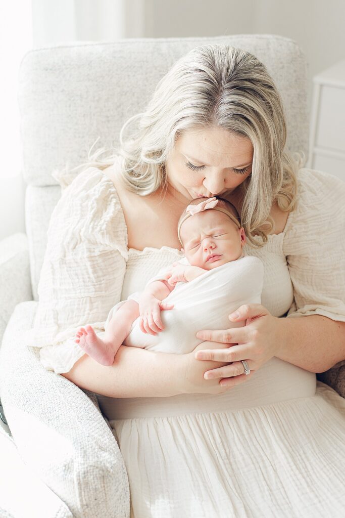 mom kissing newborn baby on forehead while sitting in neutral, light and airy nursery