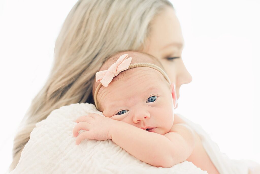 newborn baby girl is looking at camera while rested on mom's shoulder while mom's face is loking forward. 