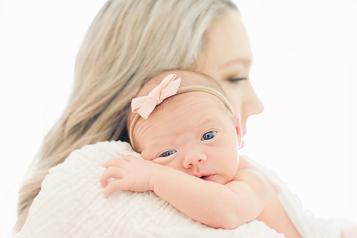 newborn baby girl is looking at camera while rested on mom's shoulder while mom's face is loking forward.