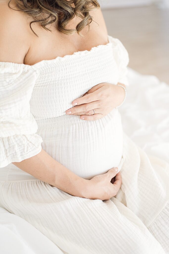 mom in white cottage off-shoulder dress with hands cradling baby for maternity session