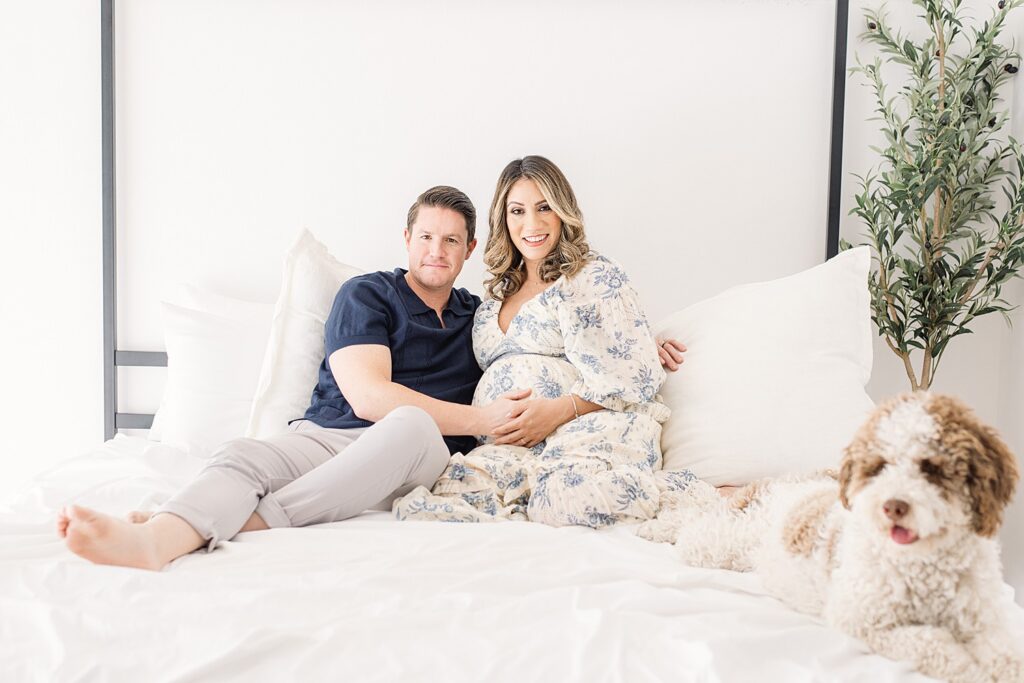 expecting parents cuddle on bed with hands on mom's baby bump. a bernadoodle is laying at their feet. 