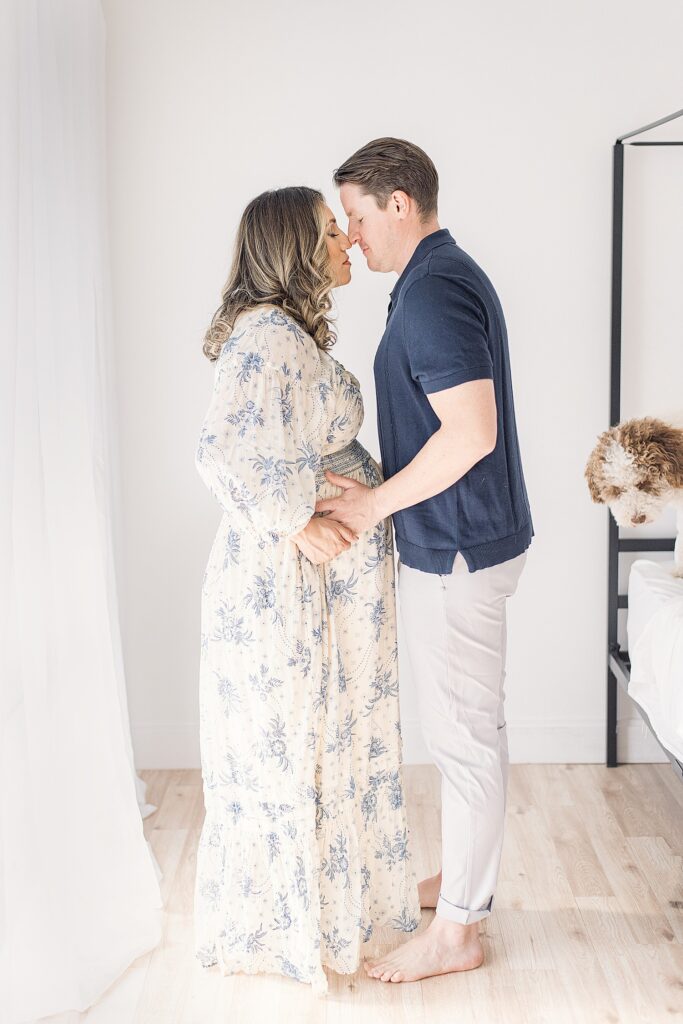 couple face to face while holding baby bump. in corner peeking is their bernadoodle