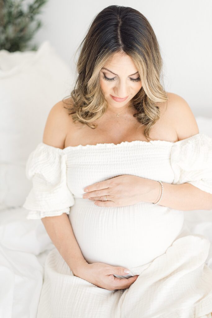 mom looking down at baby bump while cradling her belly. she's wearing a cream linen dress for studio maternity photos