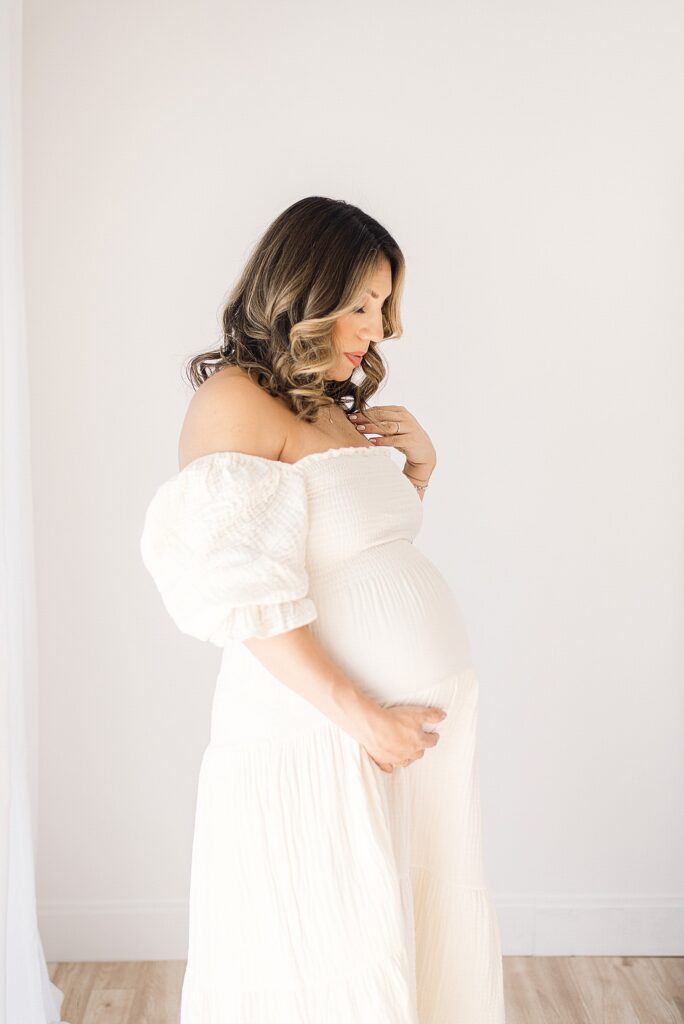 expecting mom with professional hair and makeup in cream linen dress is gazing at her baby bump for sutdio maternity photos