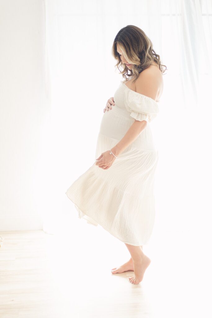 mom has one hand on baby bump while dancing and twirling flowy cream linen dress