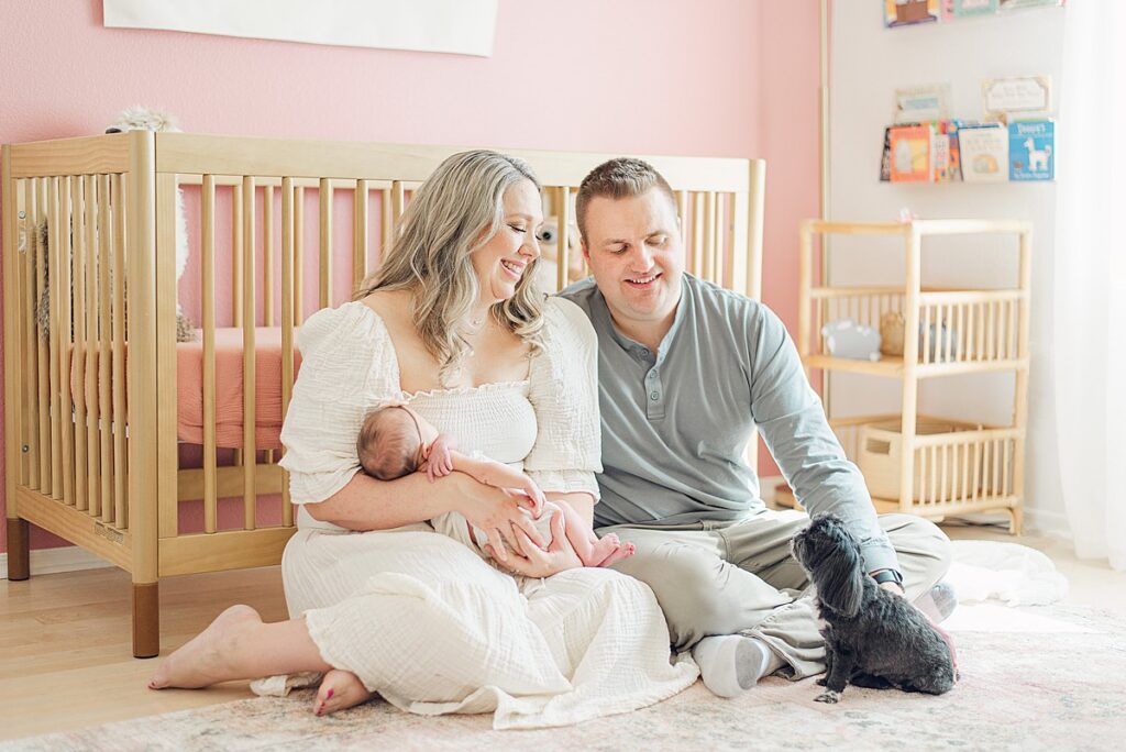 mom and dad sitting on floor of nursery in front of the crib. mom is holding baby in her arms and dad is petting dog in front of him